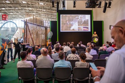 Publikum sitzt in einer Messehalle vor einer Bühne und verfolgt einen Fachvortrag mit Präsentation auf großer Leinwand.