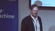 Ein Mann im Anzug spricht neben einem Bildschirm und einem Banner mit der Aufschrift 