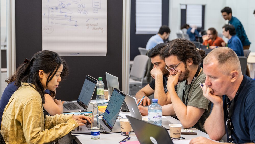 People are sitting with drinks at communal tables in an exhibition hall, engrossed in their laptops.