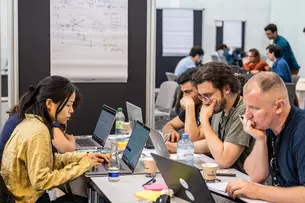 A group of people are sitting at a table in a shared workspace, concentrating on laptops, with diagrams on a blackboard behind them.