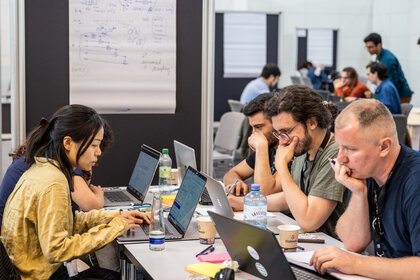 Leute sitzen mit Getränken an Gemeinschaftstischen in einer Messehalle und sind in Ihre Laptops vertieft.