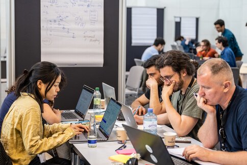 Leute sitzen mit Getränken an Gemeinschaftstischen in einer Messehalle und sind in Ihre Laptops vertieft.