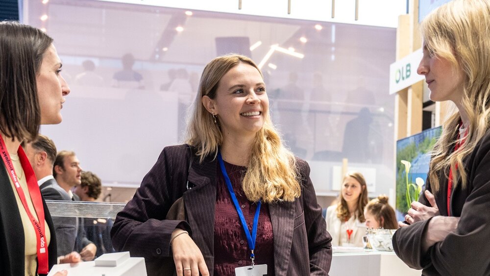 Drei Frauen in Businesskleidung stehen an einem Stand auf der EXPO REAL und unterhalten sich miteinander. Die Frau in der Mitte lächelt.
