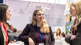 Drei Frauen in Businesskleidung stehen an einem Stand auf der EXPO REAL und unterhalten sich miteinander. Die Frau in der Mitte lächelt.