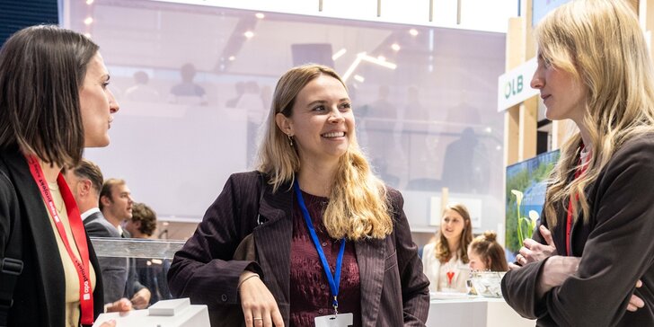 Drei Frauen in Businesskleidung stehen an einem Stand auf der EXPO REAL und unterhalten sich miteinander. Die Frau in der Mitte lächelt.