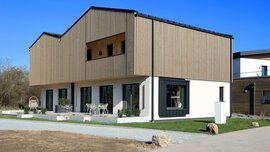 Ein modernes Haus mit senkrechten Holzlatten an der Fassade des Obergeschosses. Man sieht eine große Terrasse mit Möblierung und ein großes Fenster an der Seite.