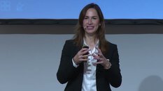 Eine Frau im schwarzen Blazer spricht in einem Headset-Mikrofon und hält einen Clicker während einer Präsentation.