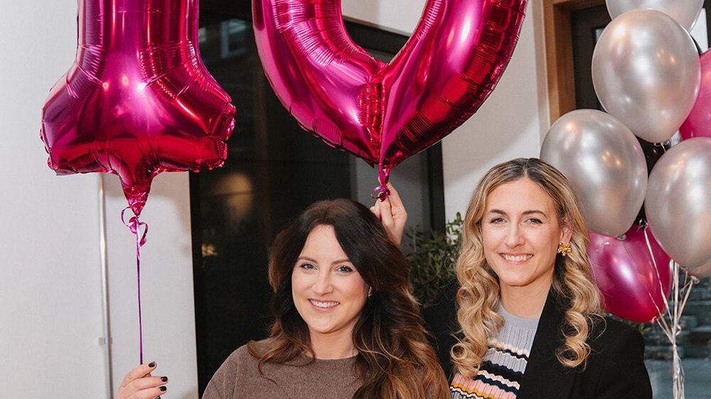 Zwei Frauen halten rosafarbene Luftballons mit der Aufschrift „10“ und feiern ein Ereignis mit silbernen und rosafarbenen Luftballons im Hintergrund