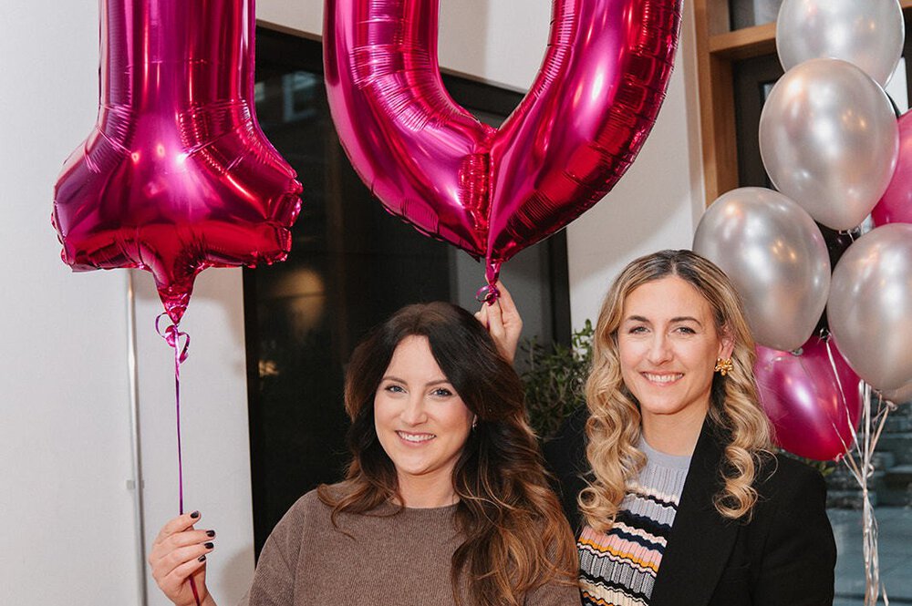 Zwei Frauen halten rosafarbene Luftballons mit der Aufschrift „10“ und feiern ein Ereignis mit silbernen und rosafarbenen Luftballons im Hintergrund