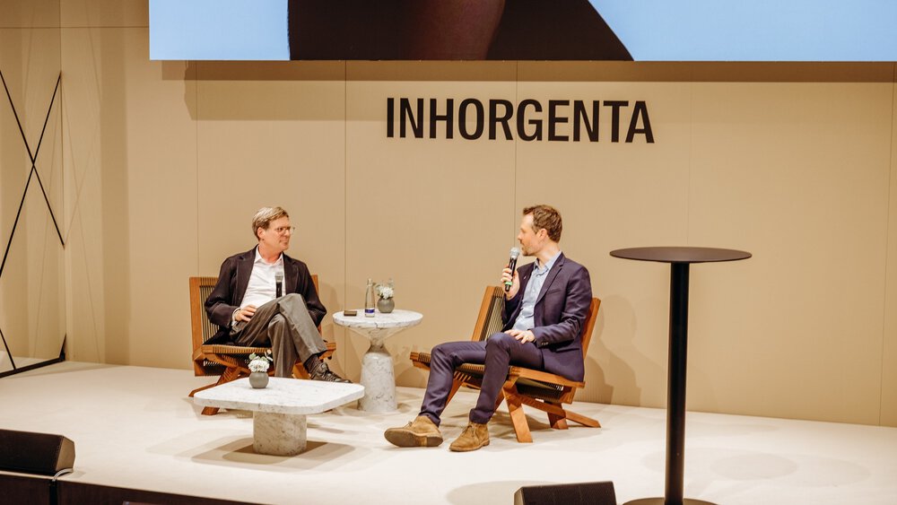 Two men with microphones are sitting on wooden chairs on a stage at INHORGENTA, chatting. Between them are two marble tables.