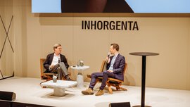 Two men with microphones are sitting on wooden chairs on a stage at INHORGENTA, chatting. Between them are two marble tables.