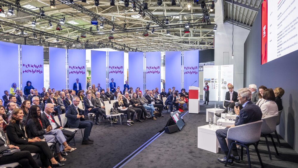 Eine Podiumsdiskussion auf der EXPO REAL in München zum Thema „Flexible Housing“. Mehrere Expertinnen und Experten sitzen auf der Bühne und sprechen vor einem großen Publikum, das aufmerksam zuhört. Im Hintergrund sind beleuchtete Wände mit dem Schriftzug „Flexible Housing“ zu sehen.