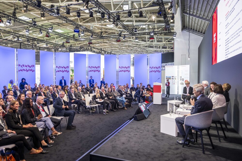 Eine Podiumsdiskussion auf der EXPO REAL in München zum Thema „Flexible Housing“. Mehrere Expertinnen und Experten sitzen auf der Bühne und sprechen vor einem großen Publikum, das aufmerksam zuhört. Im Hintergrund sind beleuchtete Wände mit dem Schriftzug „Flexible Housing“ zu sehen.