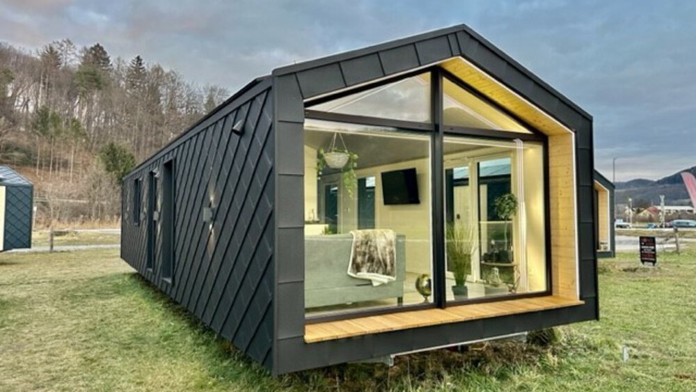 Modernes kleines Haus mit großen Fenstern, auf einer Wiese mit Bäumen und bewölktem Himmel im Hintergrund.