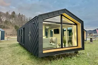 Modernes kleines Haus mit großen Fenstern, auf einer Wiese mit Bäumen und bewölktem Himmel im Hintergrund.