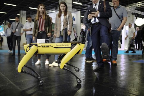 Ein gelber Roboterhund läuft vor Publikum auf einem glänzenden Boden und weicht dabei dem ausgestreckten Fuß eines Besuchers aus.