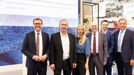A group of six people in business attire, including Hubert Aiwanger and Stefan Rummel, posing and smiling in front of an information wall for the Hydrogen Spotlight Area.