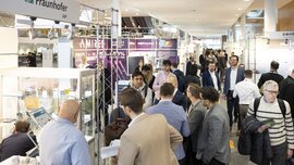 Numerous visitors walk between a row of exhibition stands at LOPEC and look at the information boards and exhibits.