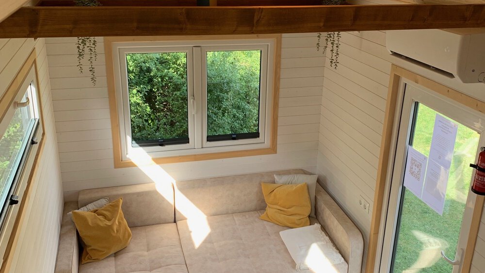 Gemütliches Zimmer mit Fensterblick ins Grüne, Balken und minimalistischer Möblierung mit gelben Kissen.