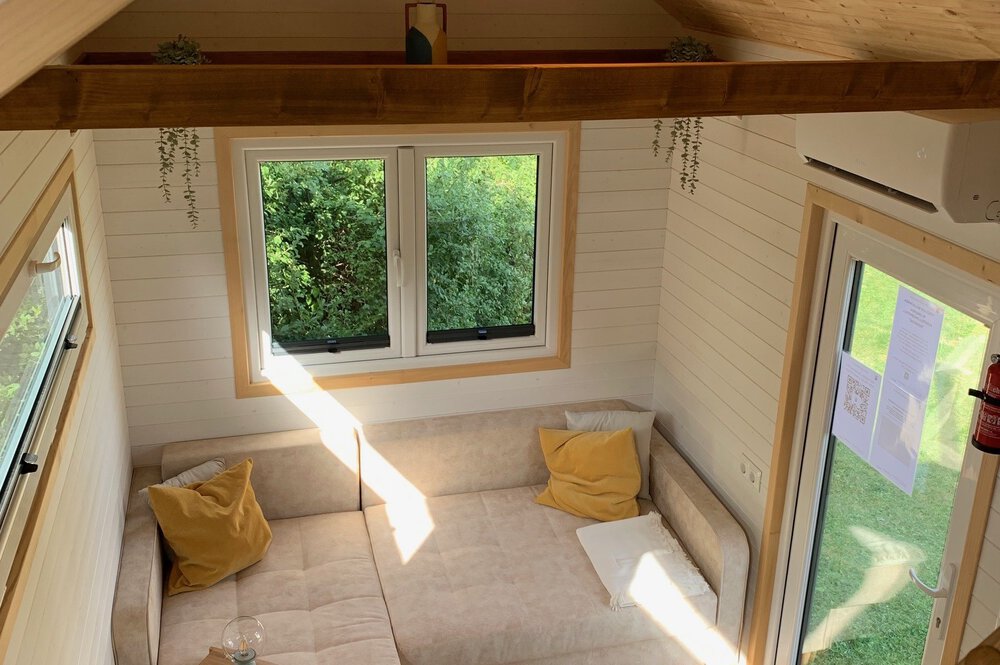 Gemütliches Zimmer mit Fensterblick ins Grüne, Balken und minimalistischer Möblierung mit gelben Kissen.