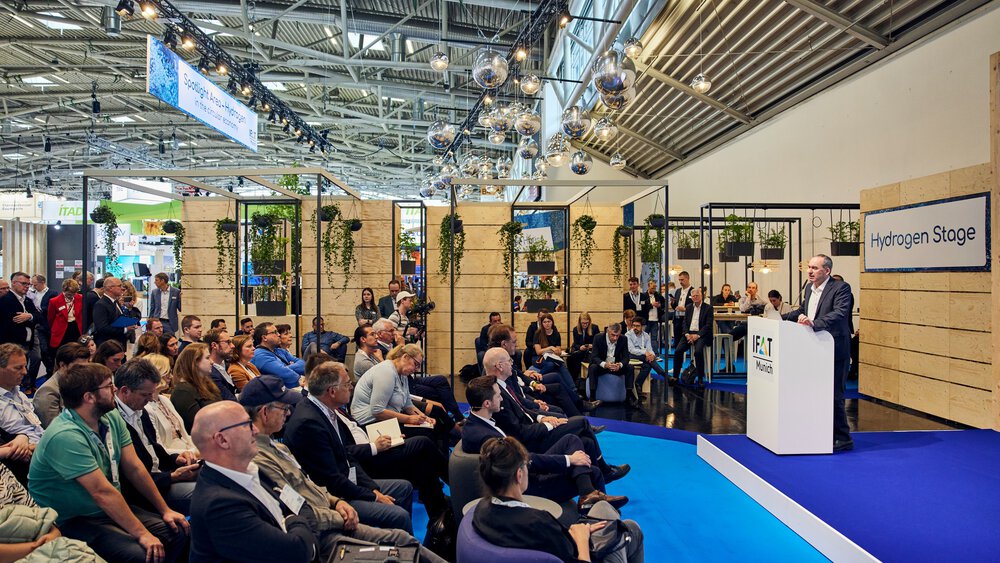 Seated audience in front of the IFAT Munich “Hydrogen Stage,” where a speaker is presenting at a podium; the blue stage area is framed by modern booth structures with hanging plants and decorative light spheres.