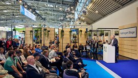 Seated audience in front of the IFAT Munich “Hydrogen Stage,” where a speaker is presenting at a podium; the blue stage area is framed by modern booth structures with hanging plants and decorative light spheres.