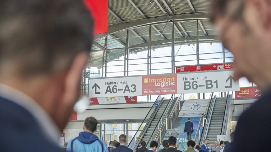 Blick zwischen zwei unscharfen Besucher im Vordergrund auf den Eingangsbereich der transport logistic mit Hinweisschildern zu den Hallen A6-A3 sowie B6-B1.