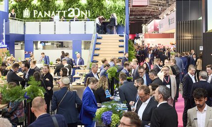 A busy exhibition booth at EXPO REAL in Munich, with numerous visitors networking, exchanging business cards, and engaging in conversations. The booth features a modern design with green plant walls, a staircase leading to an upper level, and tables with snacks and drinks. In the background, more booths and many people in a professional business atmosphere can be seen.