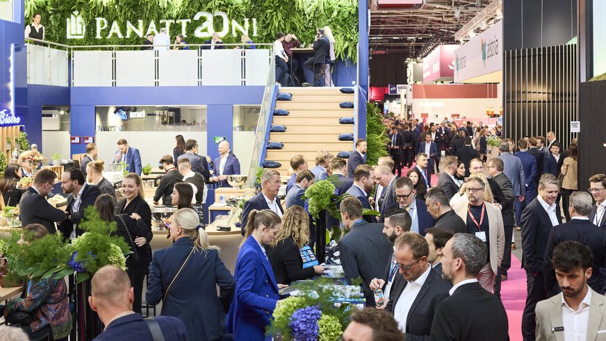 A busy exhibition booth at EXPO REAL in Munich, with numerous visitors networking, exchanging business cards, and engaging in conversations. The booth features a modern design with green plant walls, a staircase leading to an upper level, and tables with snacks and drinks. In the background, more booths and many people in a professional business atmosphere can be seen.