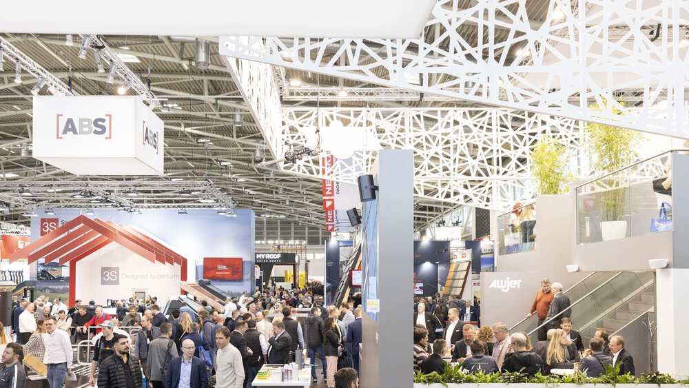 View into a hall at BAU 2025. Visitors stand around many different stands. An elaborate construction of white struts can be seen on the ceiling.