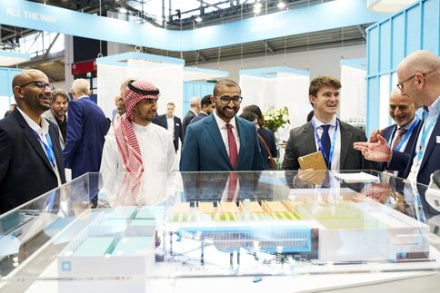 A group of businesspeople in suits and traditional clothing discuss a scale model at a trade fair booth.