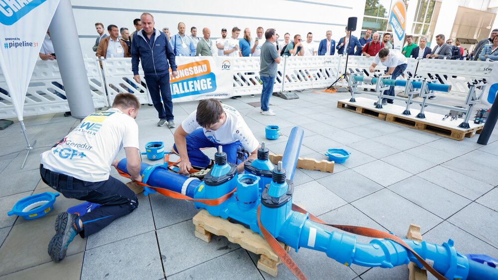 Zwei Personen montieren ein großes blaues Rohrleitungsbauteil auf Paletten bei einem Wettbewerb, im Hintergrund Zuschauer und ein Banner mit ‚Leitungsbau Challenge‘