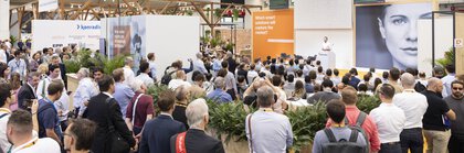 Many trade fair visitors attend a presentation on stage; a large portrait and orange signage are visible in the background.