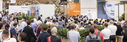 Viele Messebesucher:innen hören einem Vortrag auf einer Bühne zu; im Hintergrund ist ein großes Porträt und orangefarbene Beschilderung zu sehen.