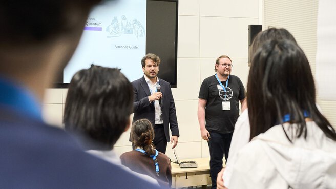 Eröffnung eines Workshops auf der World of Quantum 2025 in München: Zwei Referenten begrüßen die Teilnehmenden vor einer Präsentation zum Thema 