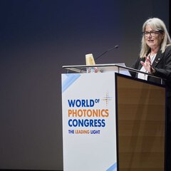 A woman with glasses, in a black suit, speaking on the podium of the World of Photonics Congress.