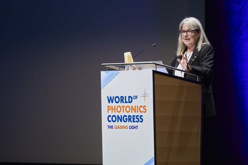 A woman with glasses, in a black suit, speaking on the podium of the World of Photonics Congress.