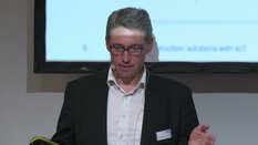 Anzugträger mit Brille spricht auf einer Bühne, nutzt ein Headset-Mikrofon und hält einen Clicker in der Hand.