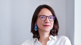 A smiling woman with shoulder-length brown hair and glasses is wearing a white blouse, dark blue trousers, blue earrings and a ring on her finger.