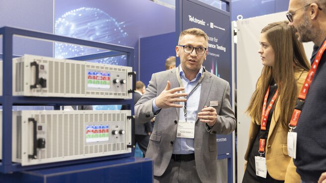 An exhibitor in a gray suit stands next to two exhibited devices in server racks and gestures to two interested trade fair visitors.