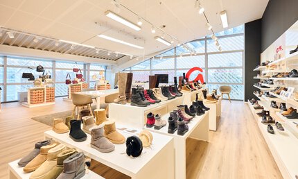 Ein großer heller Ausstellungsraum mit Glasfensterfronten und einem schrägen Dach. Auf Tischen und in Regalen werden Schuhe präsentiert.