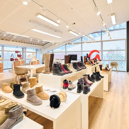 Ein großer heller Ausstellungsraum mit Glasfensterfronten und einem schrägen Dach. Auf Tischen und in Regalen werden Schuhe präsentiert.