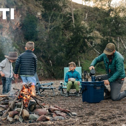 Ein Mann und drei Kinder neben einem Lagerfeuer in der Natur. Der Mann kniet vor einer blauen Box, auf der „YETI“ steht und gießt etwas in einen Becher.