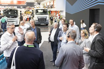 Eine Gruppe Messebesucher mit Kopfhörern lauscht einem Mann der in einer Messehalle eine Führung gibt.