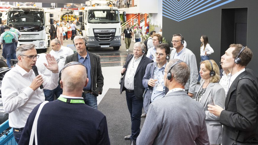 Eine Gruppe Messebesucher mit Kopfhörern lauscht einem Mann der in einer Messehalle eine Führung gibt.