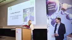 a person is standing at a lectern, in the background is a presentation with a photo of a man standing next to the stage and looking at the person at the lectern