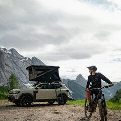 Ein Mann steht mit seinem Fahrrad vor einem Fahrzeug mit Dachzelt und Bergkulisse. Das Bild zeigt die Kombination aus Bikepacking, Dachzelt-Camping und Outdoor-Abenteuer und passt zur Podcast-Folge über Dachzelte auf der f.re.e.