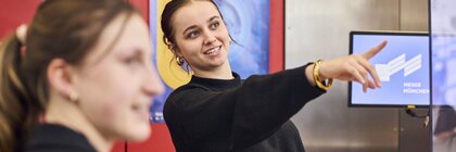 Mitarbeiterin am Information-Stand erklärt einer Besucherin etwas und zeigt nach vorn.