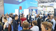 Eine Besuchergruppe mit Headsets nimmt an einer geführten Tour über die Messe teil. Ein Messevertreter mit Mikrofon erklärt die Ausstellung am Stand von SCHUNK. Im Hintergrund herrscht reger Betrieb an weiteren Ständen und Infotafeln.