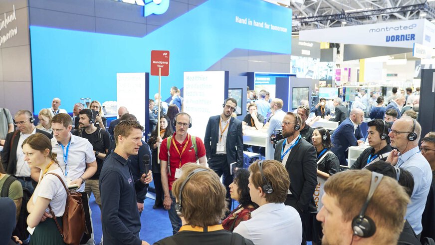 Eine Besuchergruppe mit Headsets nimmt an einer geführten Tour über die Messe teil. Ein Messevertreter mit Mikrofon erklärt die Ausstellung am Stand von SCHUNK. Im Hintergrund herrscht reger Betrieb an weiteren Ständen und Infotafeln.
